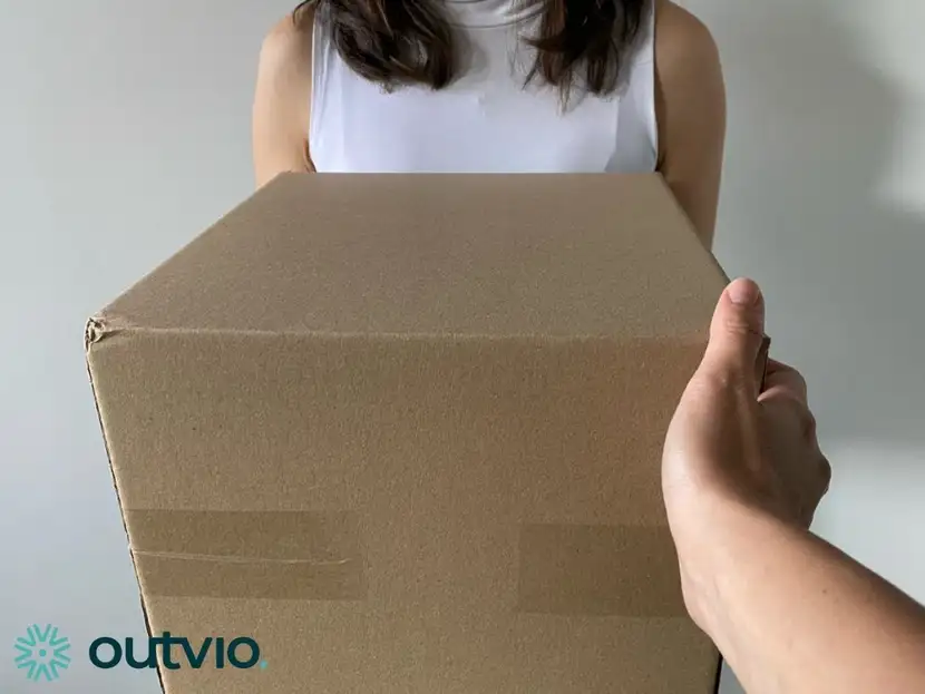 Zdjęcie przedstawiające kuriera dostarczającego paczkę do klienta który zakupił produkt za pomocą sklepu działającego na PrestaShop  kobieta ma białą bluzkę paczka tło jest szare a przesyłka kartonowa w lewym dolnym rogu napis Outvio